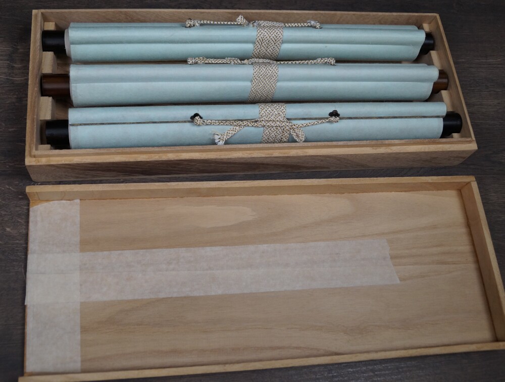 All three rolled-up triptych prints after Miyamoto Musashi with paper wrapping and cord fastening in the opened wooden box