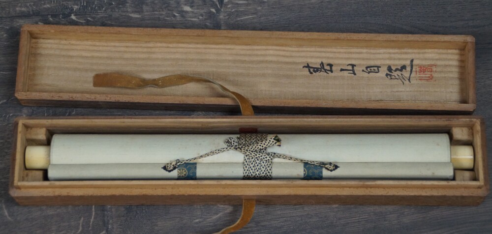 Rolled-up kakemono Encounter at the Edge of the Waterfall with ivory-colored roller ends in the opened storage box with ink inscription on the lid