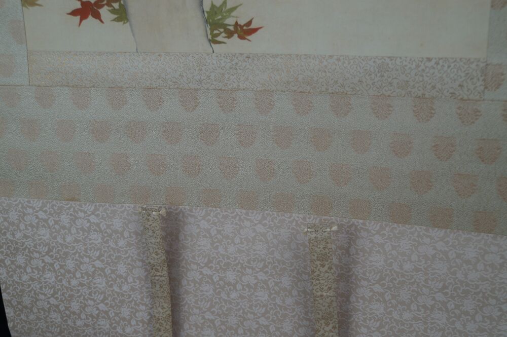 Detail view of the cream-colored patterned silk mounting with hanging cords of the hanging scroll Birds in Autumn Maple