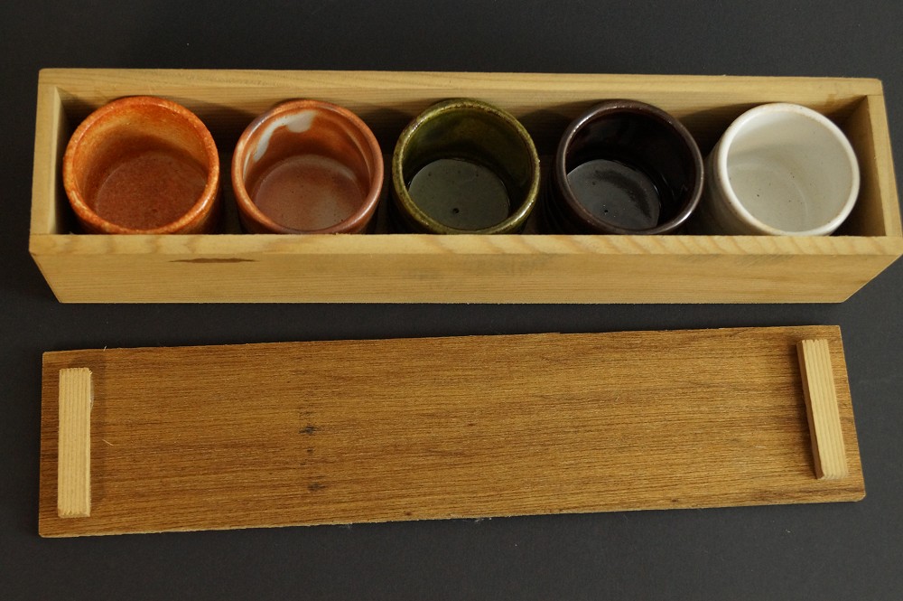 Five sake cups side by side in the elongated wooden box next to the lid set in storage box