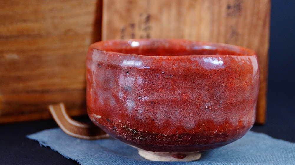 Side view of the chawan made of Raku ware by Sairi Hotoku next to the storage box on blue cloth