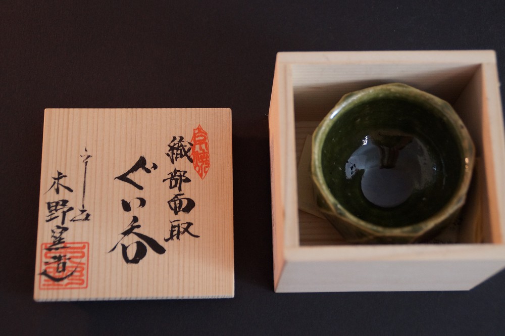 The sake cup nestled in the folding carton of the storage box next to the lid with calligraphy